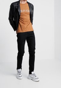 Hombre vestido con una chaqueta de cuero negra, camiseta marrón de Calvin Klein, pantalones negros y zapatillas blancas, de pie contra un fondo blanco.