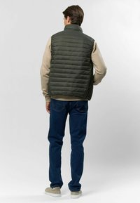 Zeleni jastučići prsluk s visokim ovratnikom, s horizontalnom qiltanom teksturom, nosi se preko svijetlosive dukserice i uparen je s tamnim trapericama.