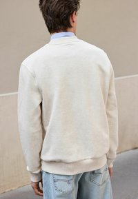 Lys beige sweatshirt med rund halsudskæring, ribbede manchetter og kant, iført over en lys blå stribet skjorte, parret med blå denim jeans.