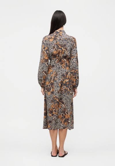 Γυναίκα με μακριά σκούρα μαλλιά που φοράει ένα μακρυμάνικο μίντι φόρεμα με floral μοτίβο σε καφέ, μαύρο και λευκό, στεκόμενη με την πλάτη προς τον θεατή.