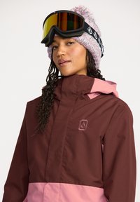 Chaqueta de esquí marrón y rosa con un cuello alto, logo bordado y tejido duradero. Combinada con un gorro de punto con pompón y gafas de esquí.