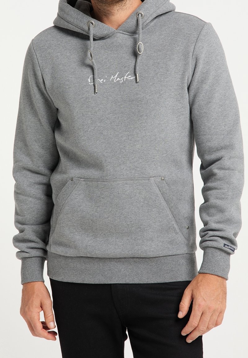 Homme portant un sweat à capuche gris avec poche avant et petit texte blanc sur la poitrine, associé à un pantalon noir.