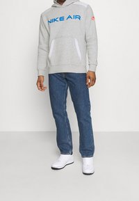 Gray hoodie with blue "NIKE AIR" text, white accents, kangaroo pocket, paired with blue denim jeans and white sneakers. Smooth materials.