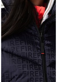 Tmavě modrá prošívaná bunda s jemným vzorem obsahujícím slovo "FIRE" v černé barvě, s kontrastní oranžovou vnitřní podšívkou a předním zipem.