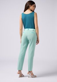 Szmaragdowa bezrękawnik w połączeniu z miętowymi spodniami w kant oraz srebrnymi sandałkami na obcasie, eksponując gładki materiał i dopasowaną sylwetkę.
