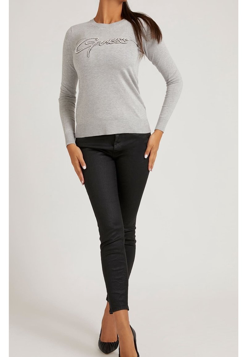 Femme portant un pull gris à manches longues avec le logo "Guess" orné de perles et un pantalon noir ajusté, debout avec une main sur la hanche et les jambes croisées.