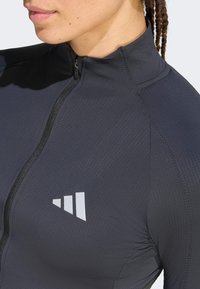 Φερμουάρ, εφαρμοστή αθλητική ζακέτα κατασκευασμένη από υφασμάτινο υλικό σε σκούρο γκρι χρώμα, με ανακλαστική λευκή λεπτομέρεια λογότυπου στο στήθος και ψηλό γιακά.