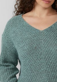 Türkis gestrickter Pullover mit V-Ausschnitt, geripptem Muster und leicht puffigen Ärmeln. Besonders hervorzuheben ist der weiche, kuschelige Stoff und die vertikale Mustergestaltung.