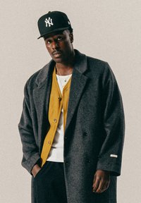 Homme portant une casquette noire des New York Yankees, un manteau oversized gris foncé, un cardigan moutarde, une chemise blanche et un pantalon noir, se tenant devant un fond uni.