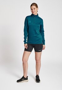 Maglietta sportiva a maniche lunghe color teal con colletto a mezza zip, abbinata a pantaloni corti compressivi neri. Presentano loghi su entrambi i capi. Sneakers nere.