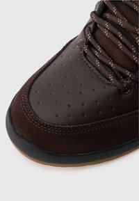 Brūna sporta apava ar gludu ādas augšdaļu un zamšādas akcentiem, kuras pirkstu daļā ir caurumi un kontrastējošas mežģīnes. Gumijas zole.