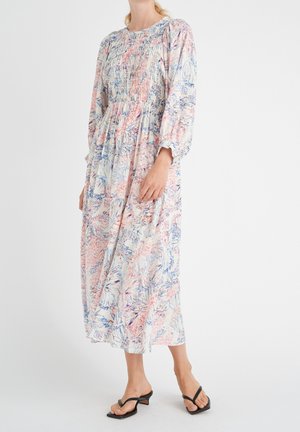 Vrouw draagt een lange, loszittende bloemenjurk met lange mouwen en een gerimpelde taille, gecombineerd met zwarte openhielhakken tegen een effen witte achtergrond.