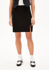 Zwarte kokerrok met een zijsplit, gemaakt van gladde stof. Gemaakt met een witte t-shirt, zwarte vest en zwart-witte schoenen.
