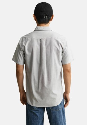 Homme portant une chemise à manches courtes gris clair avec un pli au dos, un jean bleu foncé et une casquette de baseball noire ajustable, vu de dos.