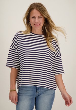 Vrouw glimlachend, met gouden creolen, een gestreept marineblauw-wit T-shirt met korte mouwen, blauwe jeans en gouden armbanden, met haar dat naar één kant waait.