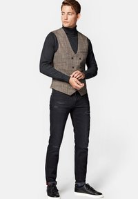 Szary kamizelka w kratę noszona na czarnym golfie, w zestawie z przetartymi czarnymi dżinsami i czarnymi trampkami. Kamizelka ma podwójne zapięcie na guziki.