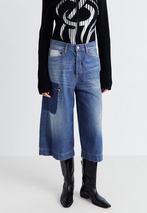 Culottes de denim en azul medio, con un bolsillo con cremallera en la parte delantera y un corte relajado. Combinados con un suéter de canalé negro de manga larga y botas negras.
