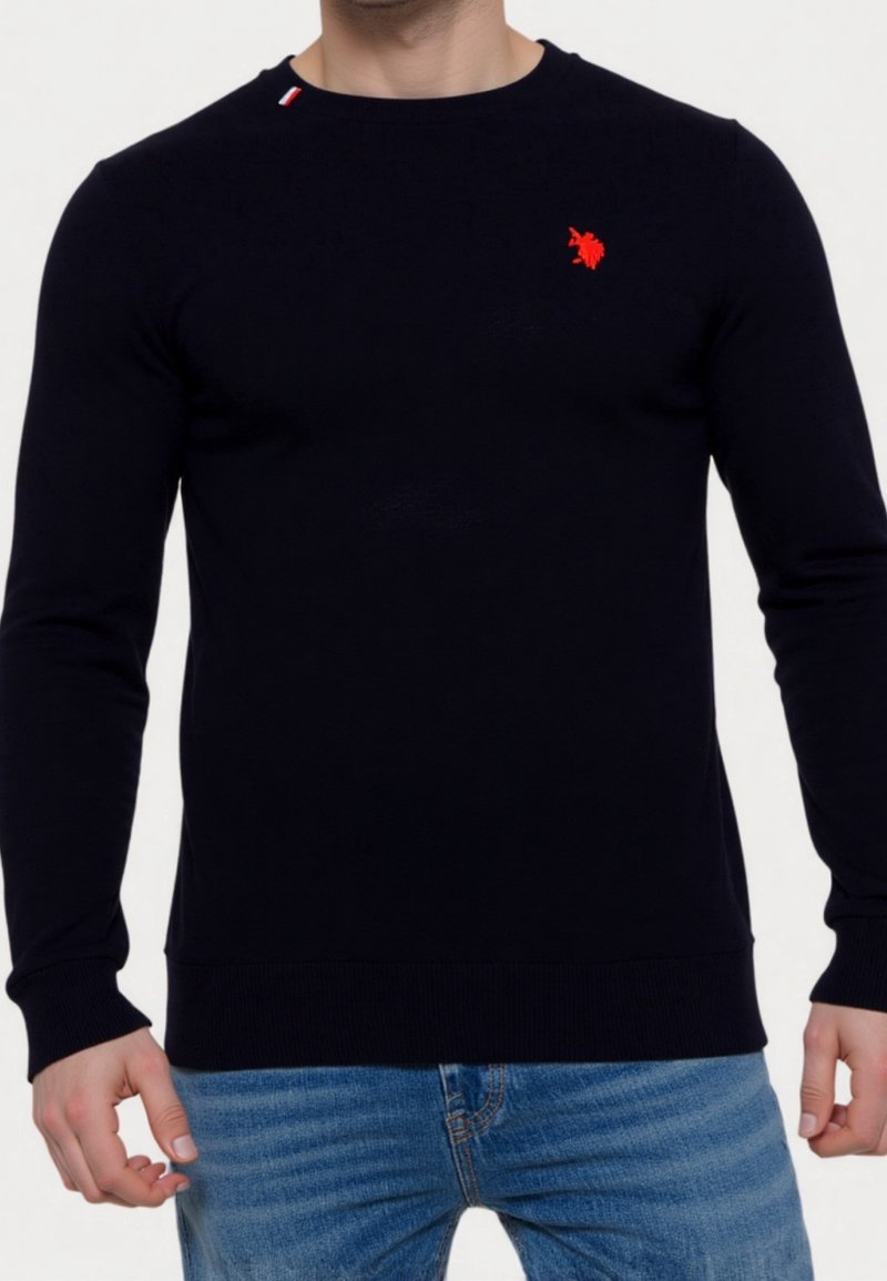 Homme portant un t-shirt noir uni à manches longues avec un petit logo rouge sur la poitrine et un jean bleu clair sur fond blanc.