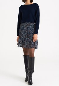 Granatowy sweter z prążkowanym wzorem w połączeniu z wzorzystą spódnicą w ciemnoniebieskim kolorze z kwiatowymi detalami, falbaną u dołu oraz kozakiem do kolan w kolorze czarnym.