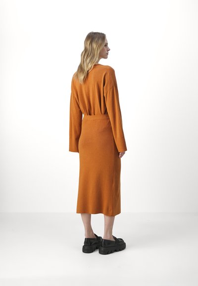 Robe longue à manches longues couleur orange brûlé avec une coupe décontractée et une taille nouée. Comprend un ourlet mi-long et un tissu texturé. Portée avec des chaussures noires à semelle épaisse.