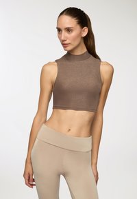 Top corto senza maniche marrone in un materiale brillante e elasticizzato con collo alto, abbinato a leggings beige a vita alta.