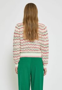 Strickpullover mit wellenförmigen roten und grünen Streifen auf einem cremefarbenen Hintergrund, mit gerippten Bündchen und Saum, kombiniert mit grünen, weiten Hosen.