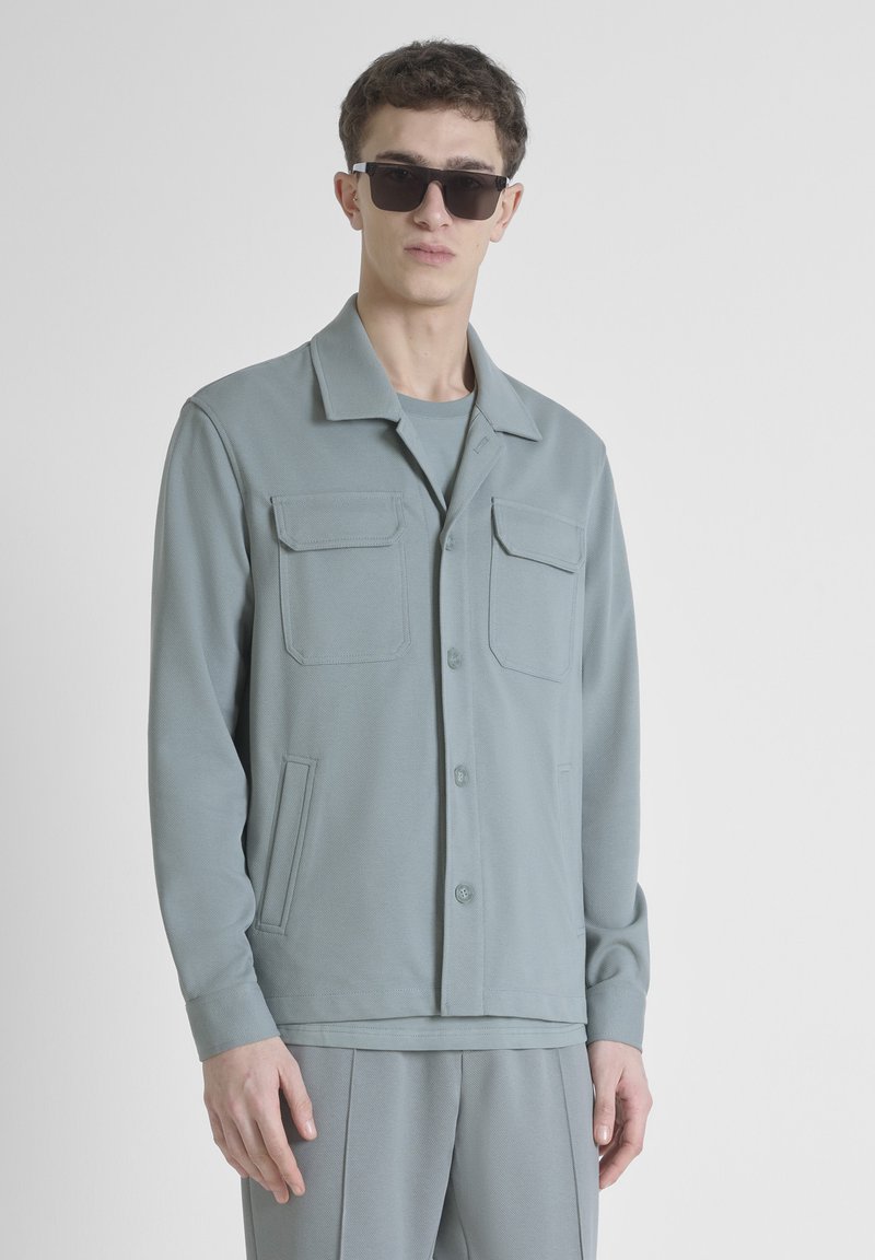 Giacca grigia chiara con colletto, due tasche sul petto e tasche laterali. Realizzata in un tessuto morbido, abbinata a una camicia e pantaloni coordinati.