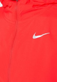 Nike vycházková bunda na zip v jasně červené barvě. Vyrobena z texturované tkaniny, na levé části hrudi se nachází stříbrné logo Nike.
