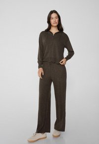 Brązowa, teksturowana kurtka zapinana na zamek z regulowanym sznurkiem oraz dopasowane, luźne spodnie. Model nosi jasne sneakersy. Prosty, casualowy design stroju.