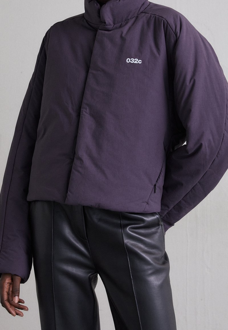 Personne portant une doudoune violet foncé avec le logo "032c" sur la poitrine et un pantalon en cuir noir sur un fond clair uni.