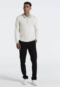 Cremefarbener Poloshirt mit langen Ärmeln und schwarzem gestreiften Kragen, kombiniert mit schwarzen Hosen und weißen Sneakers. Einfaches Design und taillierte Silhouette.