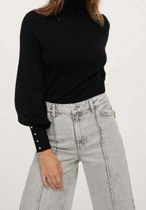 Femme portant un col roulé noir à manches longues avec des boutons en perle sur les poignets et un jean large taille haute gris clair.