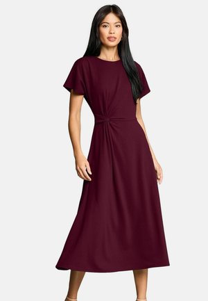 Frau, die ein knielanges, kurzärmeliges dunkelrotes Kleid mit seitlichem Knotendetail trägt, steht vor einem einfarbigen hellen Hintergrund.