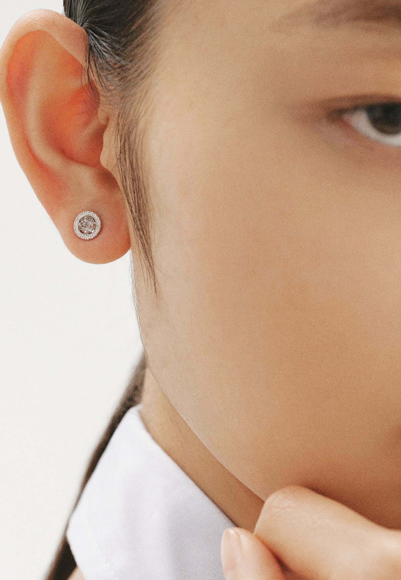Pendientes de plata con una gema redonda y clara, engastada en un contorno circular texturizado, mostrados en un primer plano de una oreja.