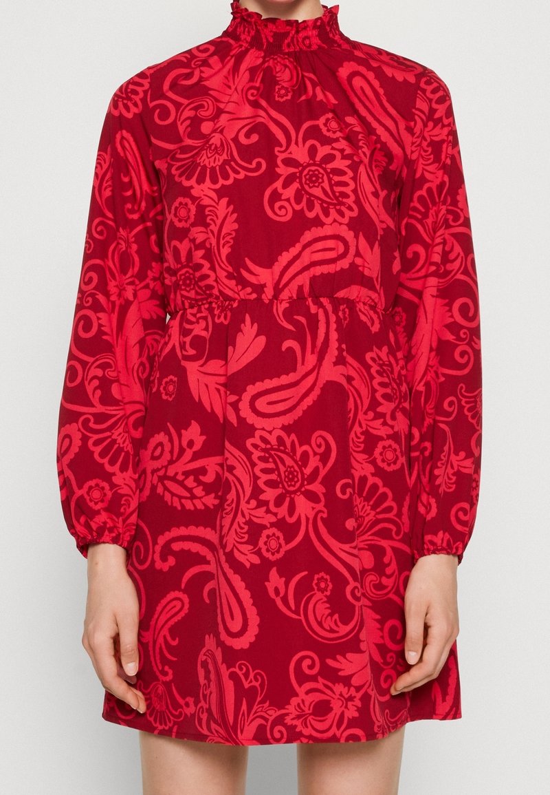 Robe rouge à manches longues avec col haut à volants et motif floral paisley, portée par une personne montrée du cou jusqu'à mi-cuisse.