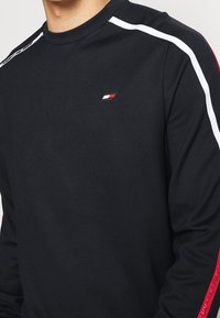 Maglietta nera a maniche lunghe con collo tondo, con strisce bianche laterali e un piccolo logo sul petto. Tessuto morbido e design aderente.