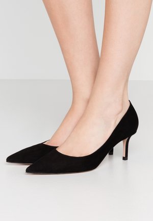 Stopy w czarnych zamszowych butach na niskim obcasie typu kitten heel z ostrym noskiem, na jasnoszarej podłodze, pokazujące skórę kostki i dolnej części nogi.