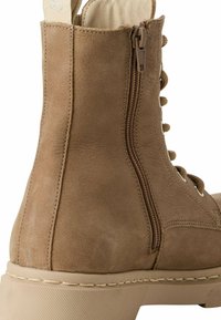 Tan suede ankle boot with a textured surface, side zipper, and lace-up front. Chunky sole with stitching detail, designed for comfort.