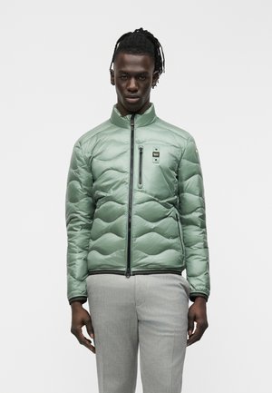 Muž oblečený v svetlozelenom prešívanom kabáte so zipsom vpredu a svetlosivých nohaviciach, stojaci na bielom pozadí.