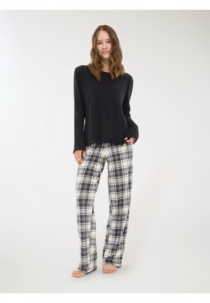 Top nero a maniche lunghe con orlo sfrangiato, abbinato a pantaloni pigiama a quadri bianchi, grigi e blu navy. Modella in piedi a piedi nudi contro uno sfondo chiaro.
