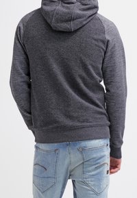 Homme portant un sweat à capuche gris foncé et un jean bleu clair, vu de dos sur un fond blanc uni.
