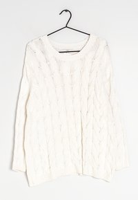 Un pull en tricot blanc avec un motif en relief, un col rond, des manches longues et des poignets et un ourlet côtelés, suspendu à un crochet noir.