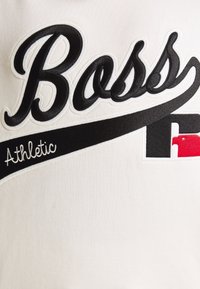 Tissu blanc avec texte brodé en noir "Boss" et "Athletic", agrémenté d'une bande d'accent noir et rouge. Texture lisse avec lettrage en relief.