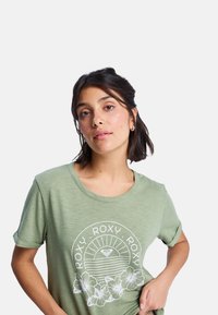 T-shirt en coton vert clair avec col rond et manches retroussées. Présente un imprimé graphique blanc de soleil et de fleurs avec le texte "ROXY".