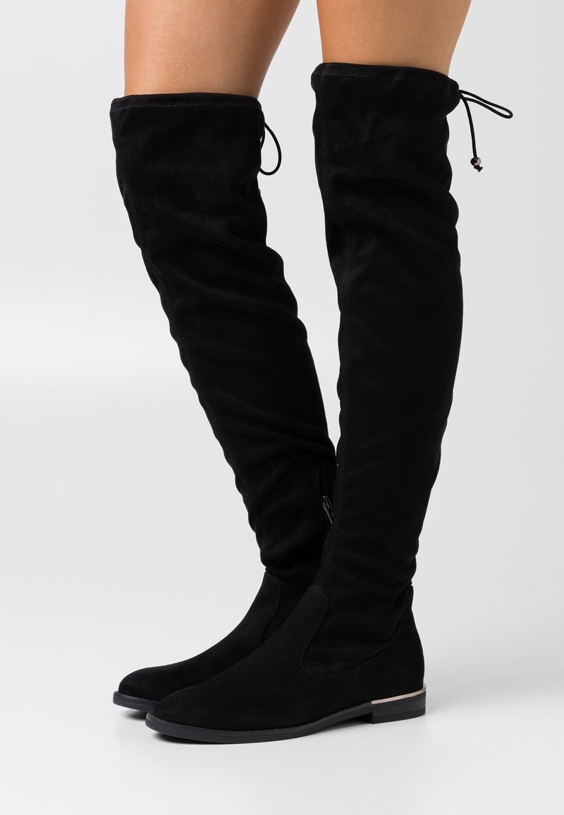 Bottes plates noires au-dessus du genou avec bouts arrondis, texture suédée et lien réglable à l’arrière, portées sur jambes nues devant un fond uni.