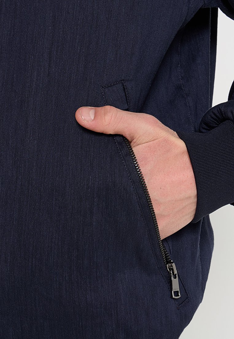 Marinejacke mit strukturiertem Finish, Reißverschlusstasche und gerippten Bündchen. Die Hand ruht auf einer seitlichen Tasche mit Reißverschlussdetails.