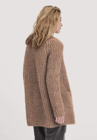 Cardigan en maille marron avec une coupe décontractée et une texture côtelée, doté de manches longues et d'une poche sur le devant.