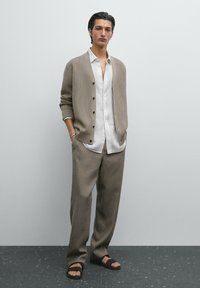 Beige gerippter Cardigan, weißes Hemd mit Knöpfen, hellbraune gestreifte Hose und dunkelbraune Sandalen. Glatte Texturen mit kontrastierenden Farben.