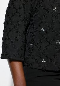 Schwarze, strukturierte Bluse mit 3/4-Ärmeln, die mit floralen Applikationen und klaren Perlenakzenten verziert ist. Der Stoff hat ein dezentes, erhabenes Muster.