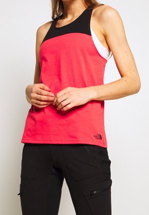 Femme portant un haut sans manches rouge et noir avec une couche blanche en dessous et un pantalon cargo noir, mains croisées devant la taille.